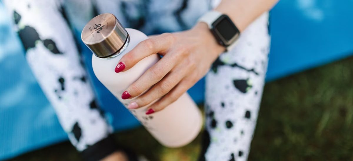 Crop unrecognizable female wearing sports leggings smart watch and holding bottle with water while relaxing after training on mat outside
